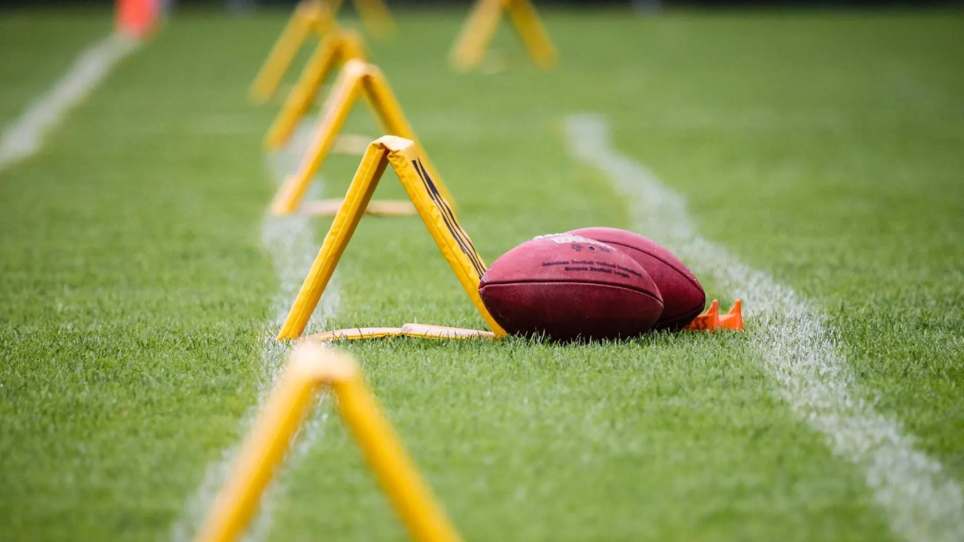 Two American footballs, one resting on an orange kicking tee, sitting on a white yard line of a green grass field, with a row of yellow triangular agility hurdles trailing into the background.