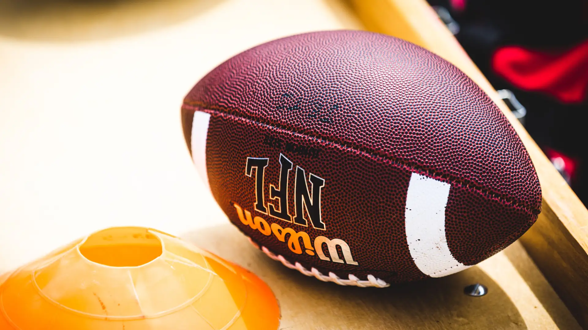 A close-up of a Wilson NFL American football showing its pebbled leather texture and white stripes, resting on a wooden surface next to an orange plastic practice cone.