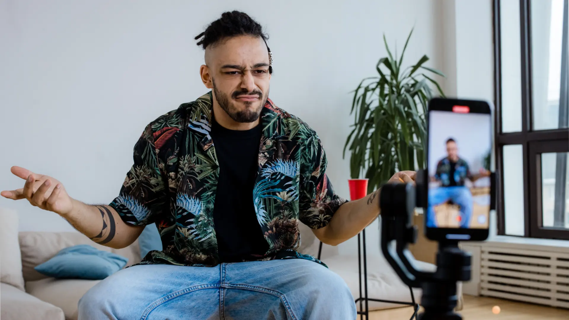 A young male content creator sitting on a couch gesturing with open hands while filming a reaction video on a smartphone mounted on a gimbal stabilizer.