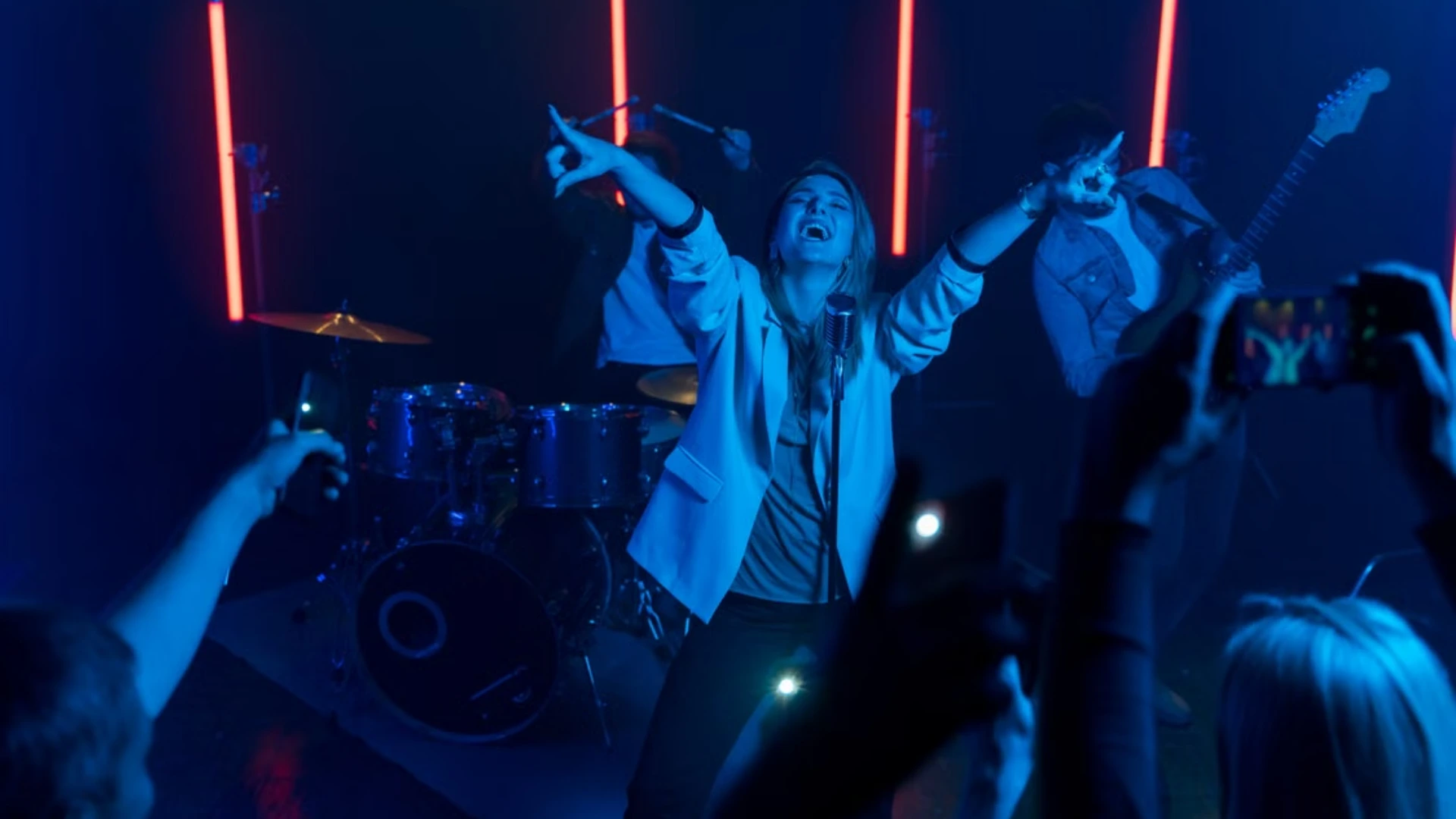A female lead singer performing with arms raised into a vintage microphone alongside a live band, illuminated by blue stage lighting and vertical red neon tubes while audience members record with smartphones.