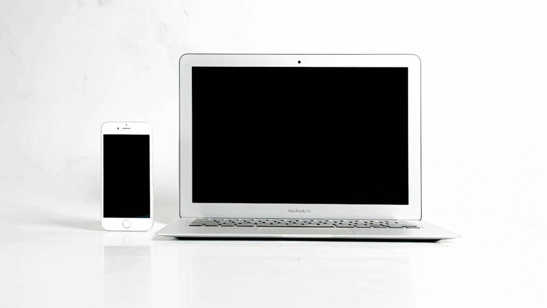 A silver laptop and white smartphone mockup with blank black screens standing on a reflective white background for responsive video template previews.